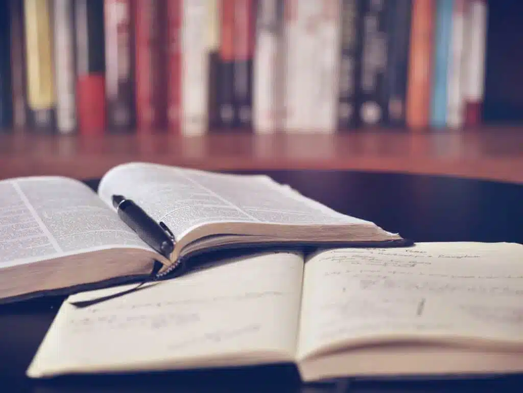 aaron-burden-QJDzYT_K8Xg-unsplash An open book and a notebook with a pen on a table, with a bookshelf in the background.